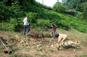 Three individuals performing archaeological work with shovels and other tools. Image of hill with dirt and overgrown foliage behind them. 